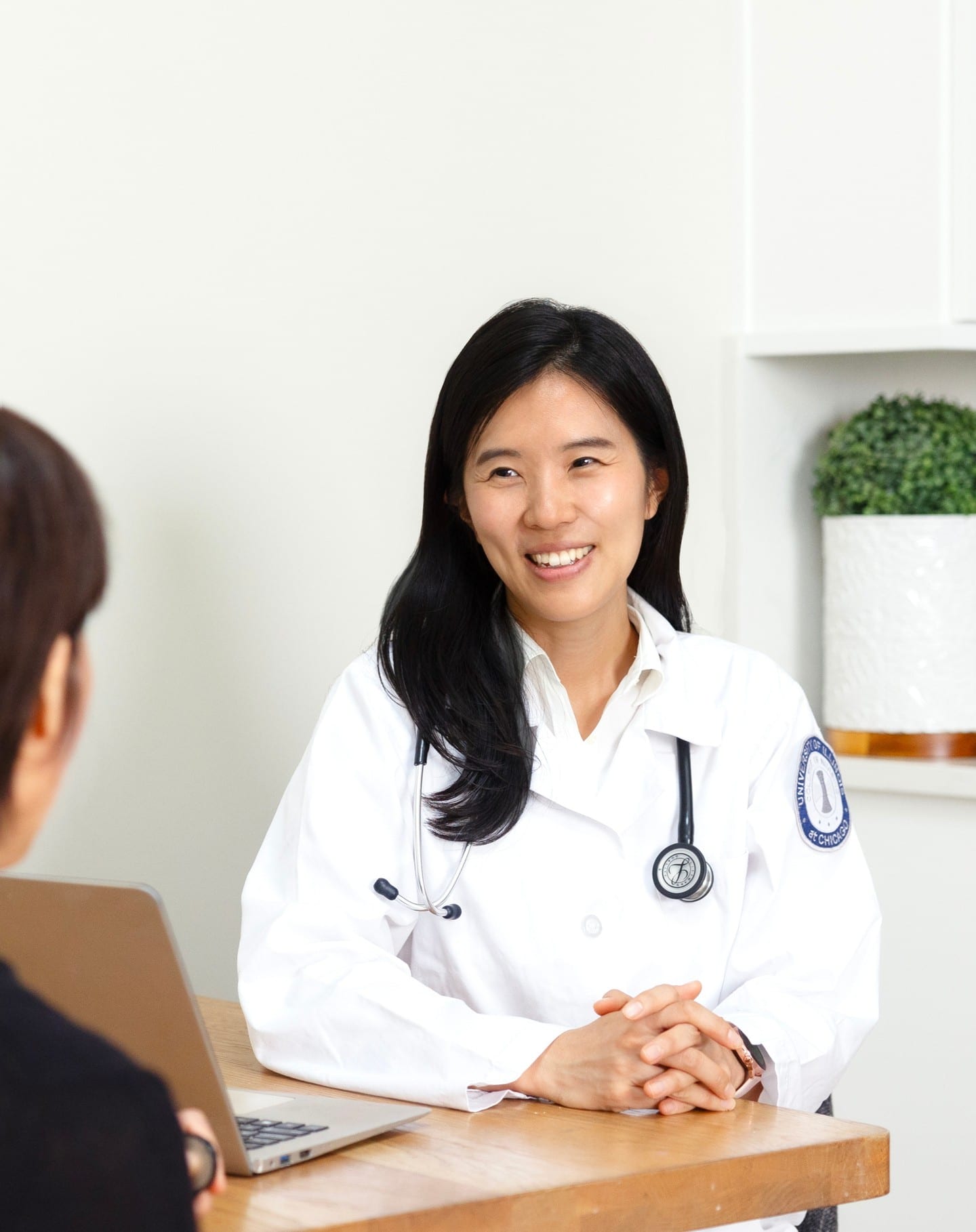 Physician in a UIC College of Medicine white coat and stethoscope smiling across a desk at a colleague with a laptop