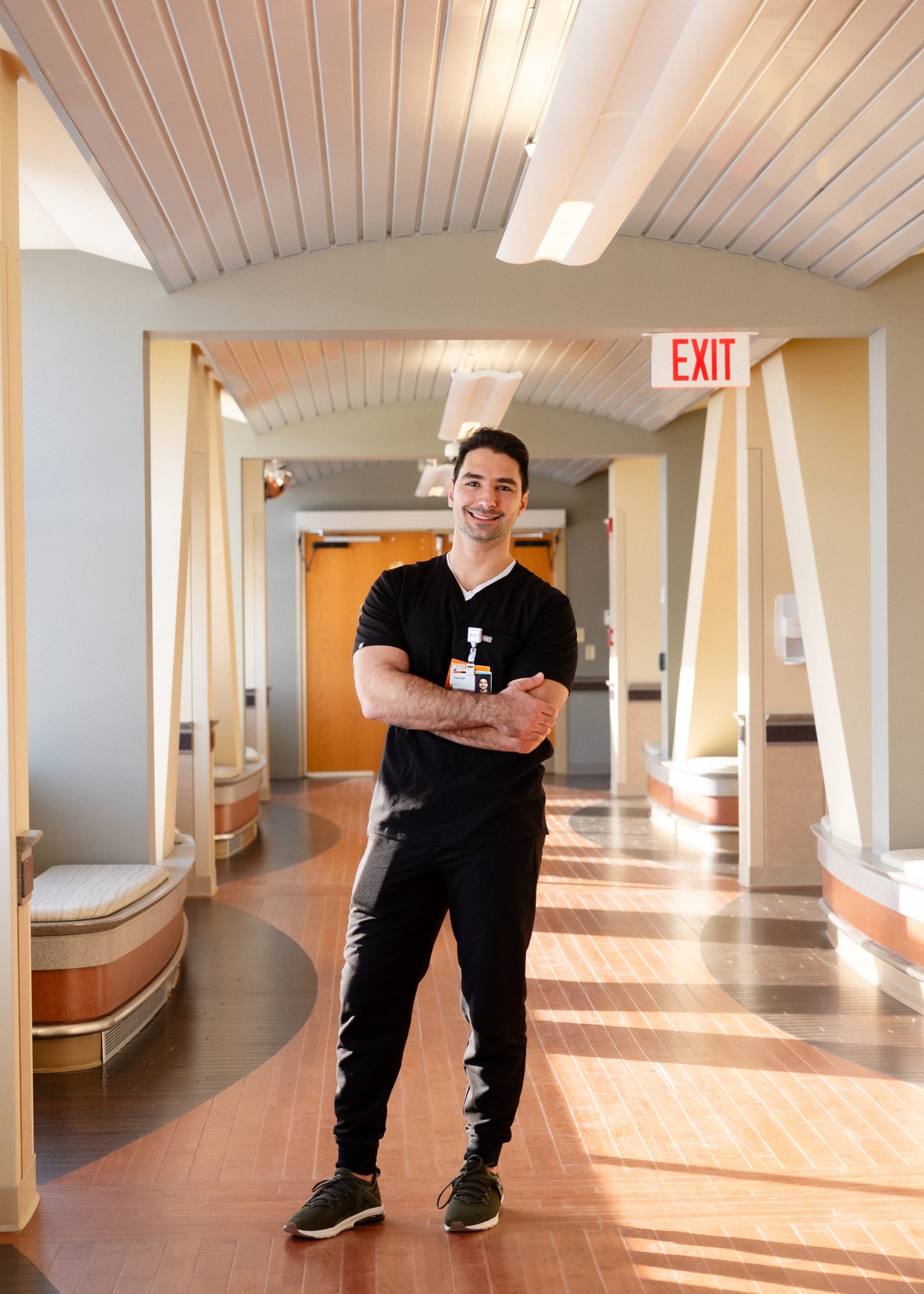 Clinician in black scrubs, arms crossed, standing in a sunlit hospital corridor