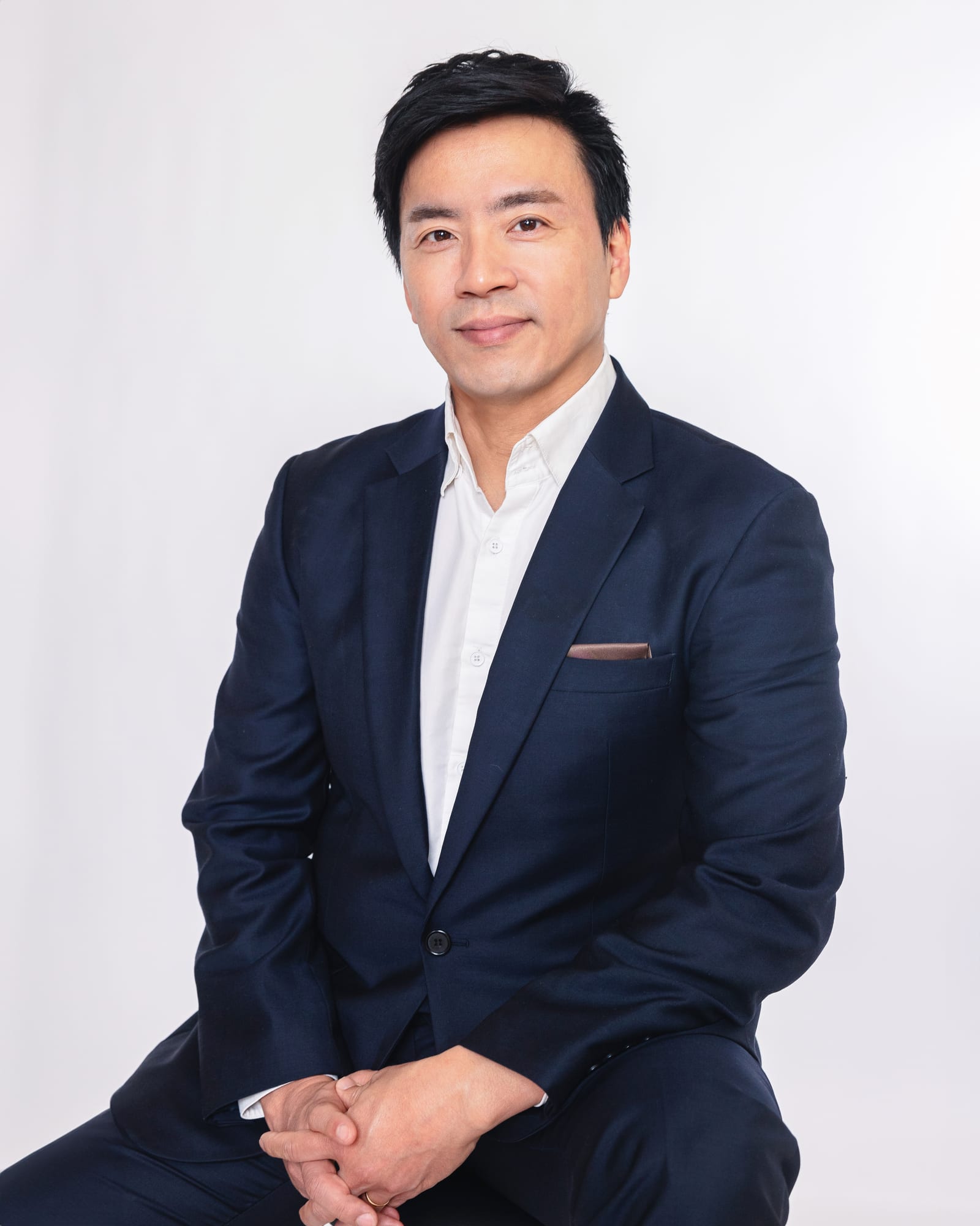 Professional in a navy suit and open white shirt seated with hands clasped, smiling softly against a white studio backdrop