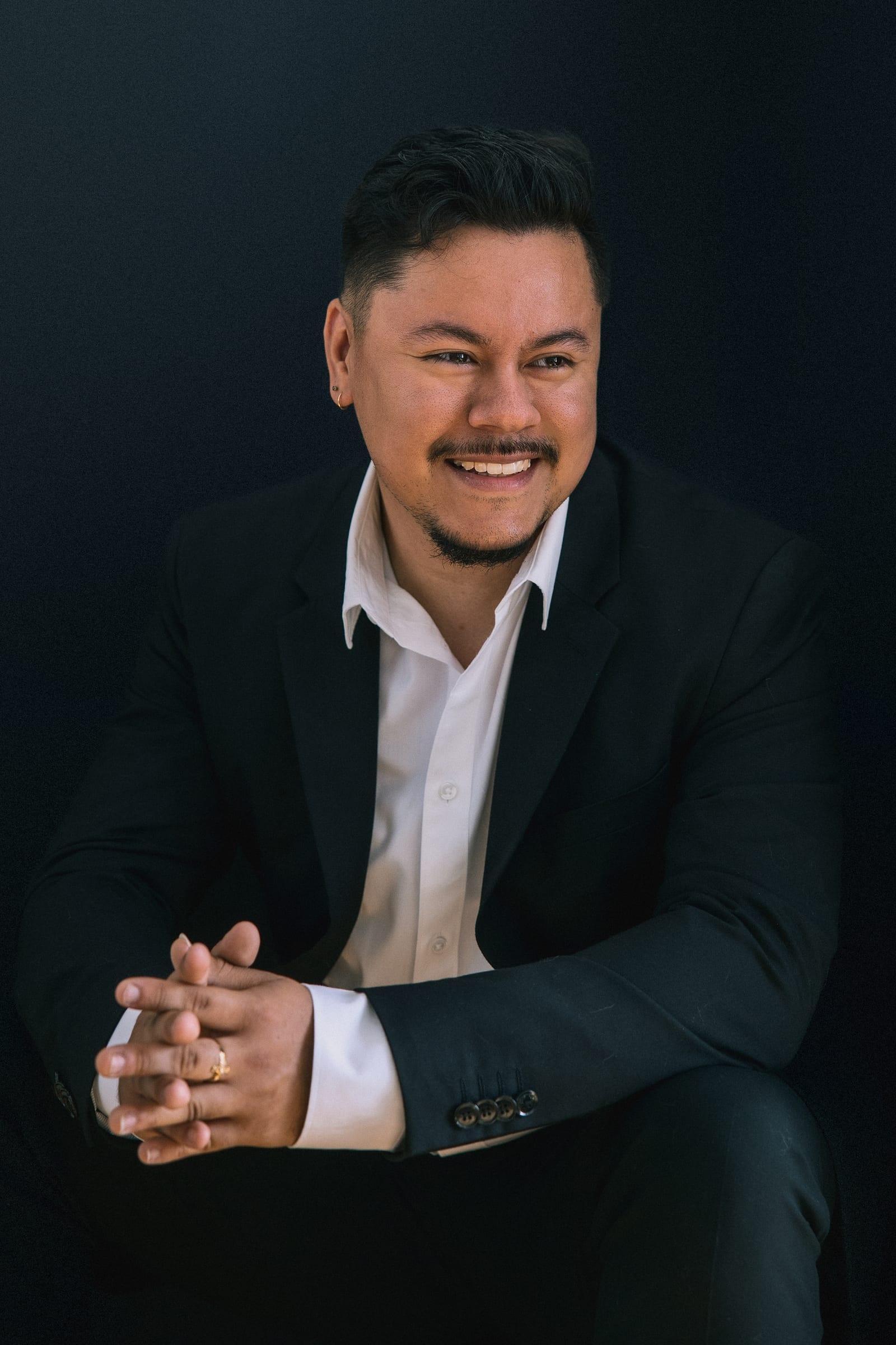 Executive in a black jacket and open white shirt, hands clasped, smiling against a charcoal ground
