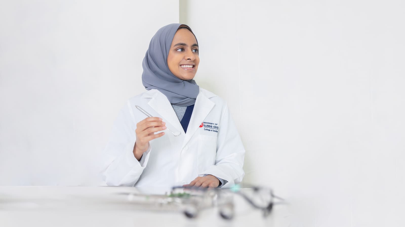 Physician wearing a hijab and white coat, seated at a bright desk beside a pair of glasses