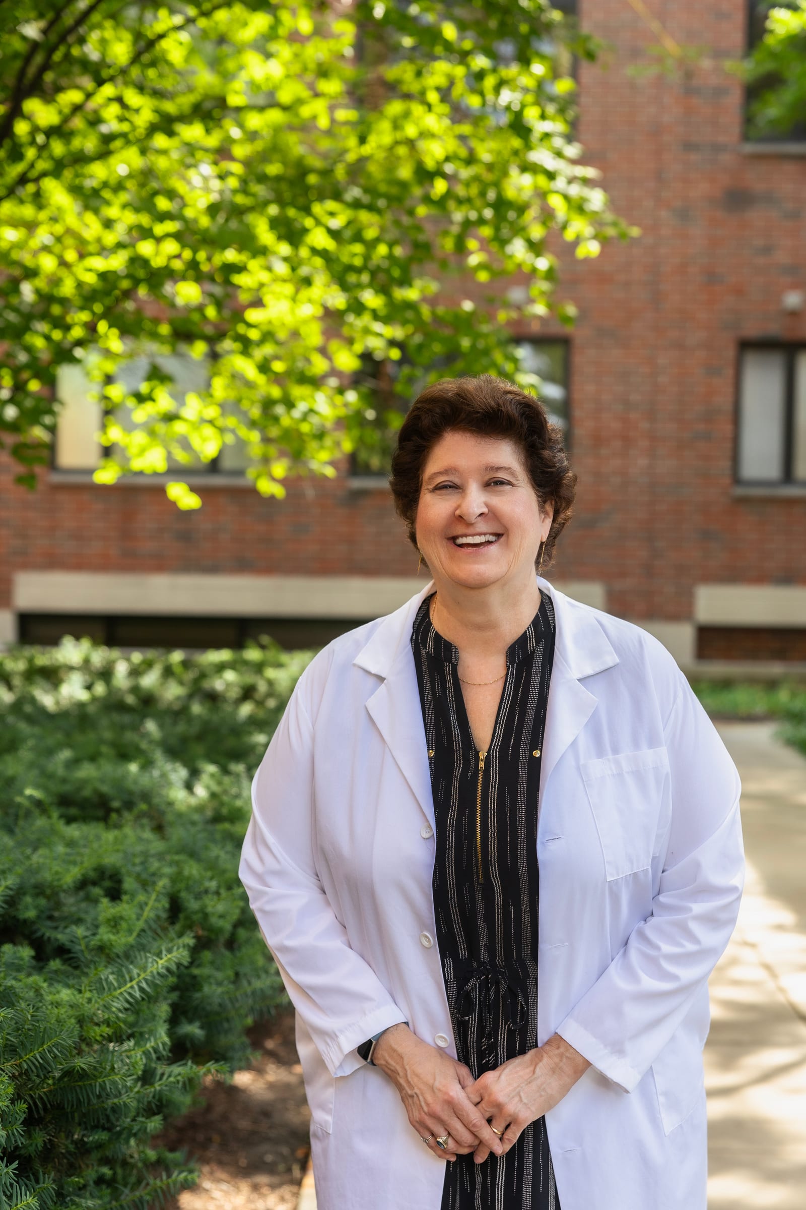 Woman physician in a white coat laughing, hands clasped, outside a red-brick building with sunlit trees behind her