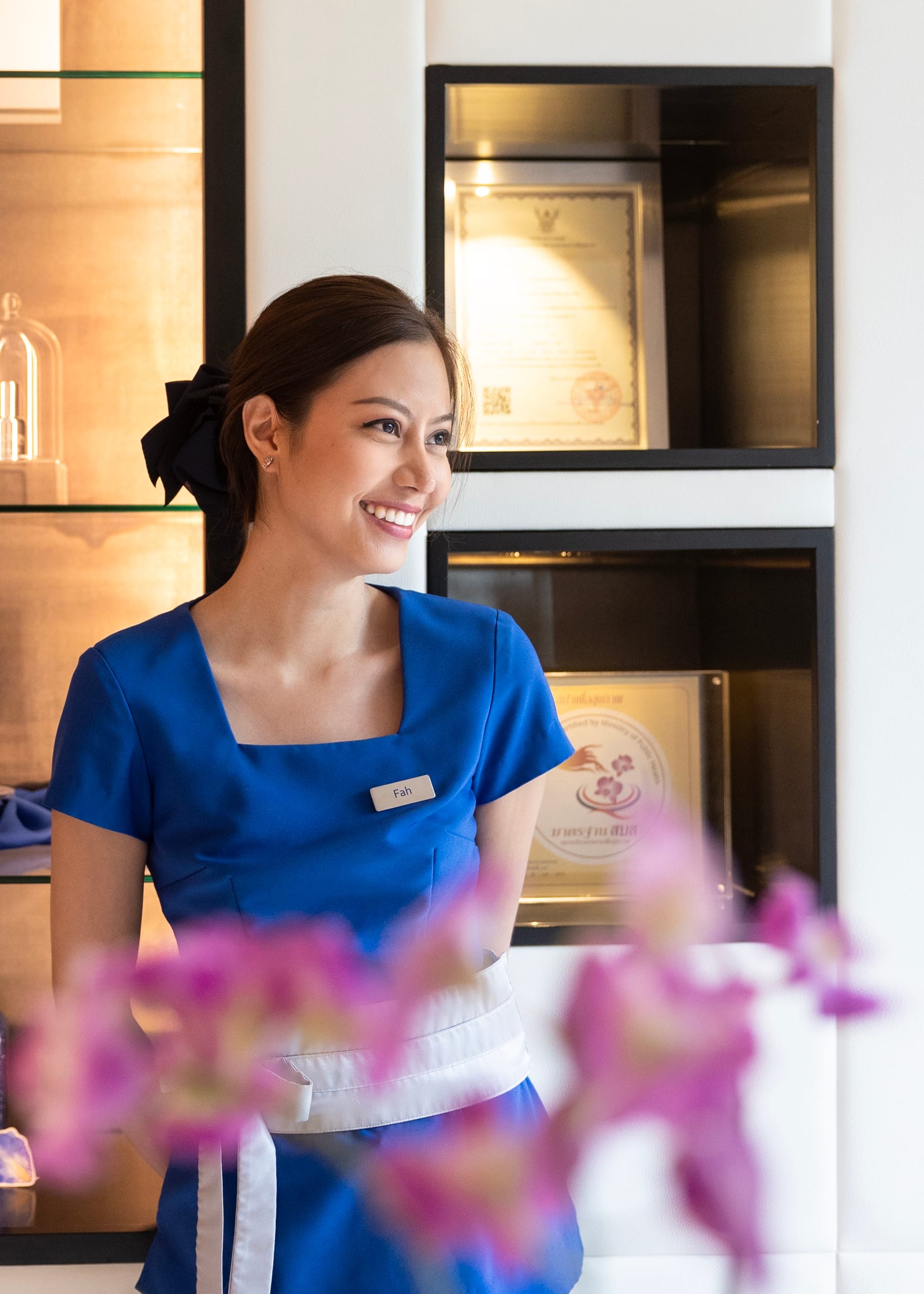 Practitioner in a blue uniform smiling in a bright treatment suite, orchids in the foreground