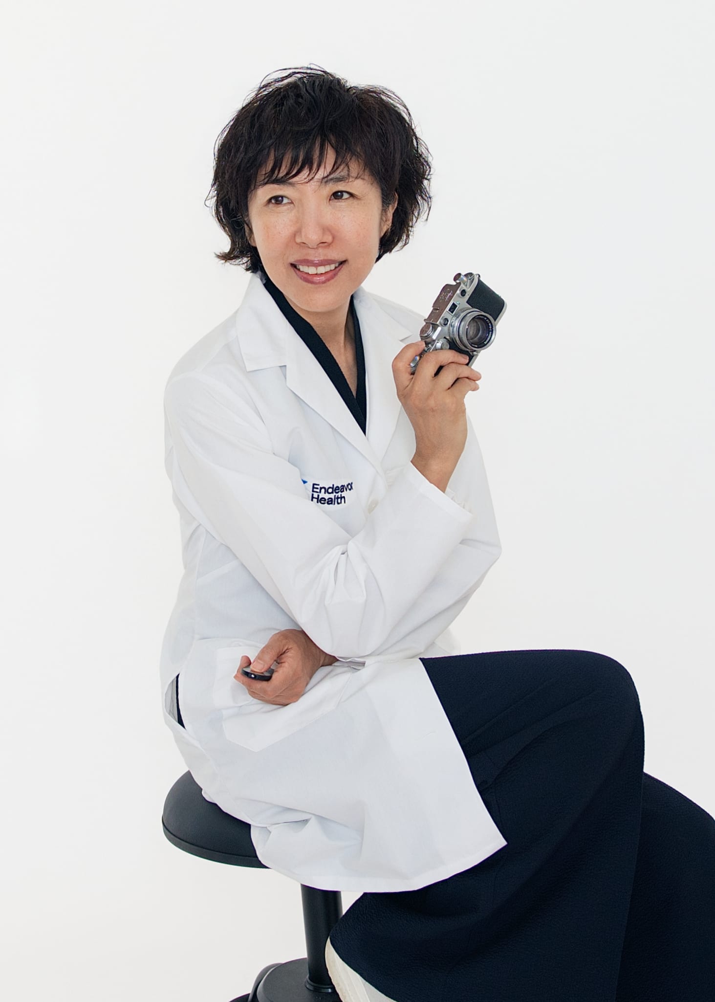 Kay Choi seated in a hospital white coat, holding a rangefinder camera and looking to the lens