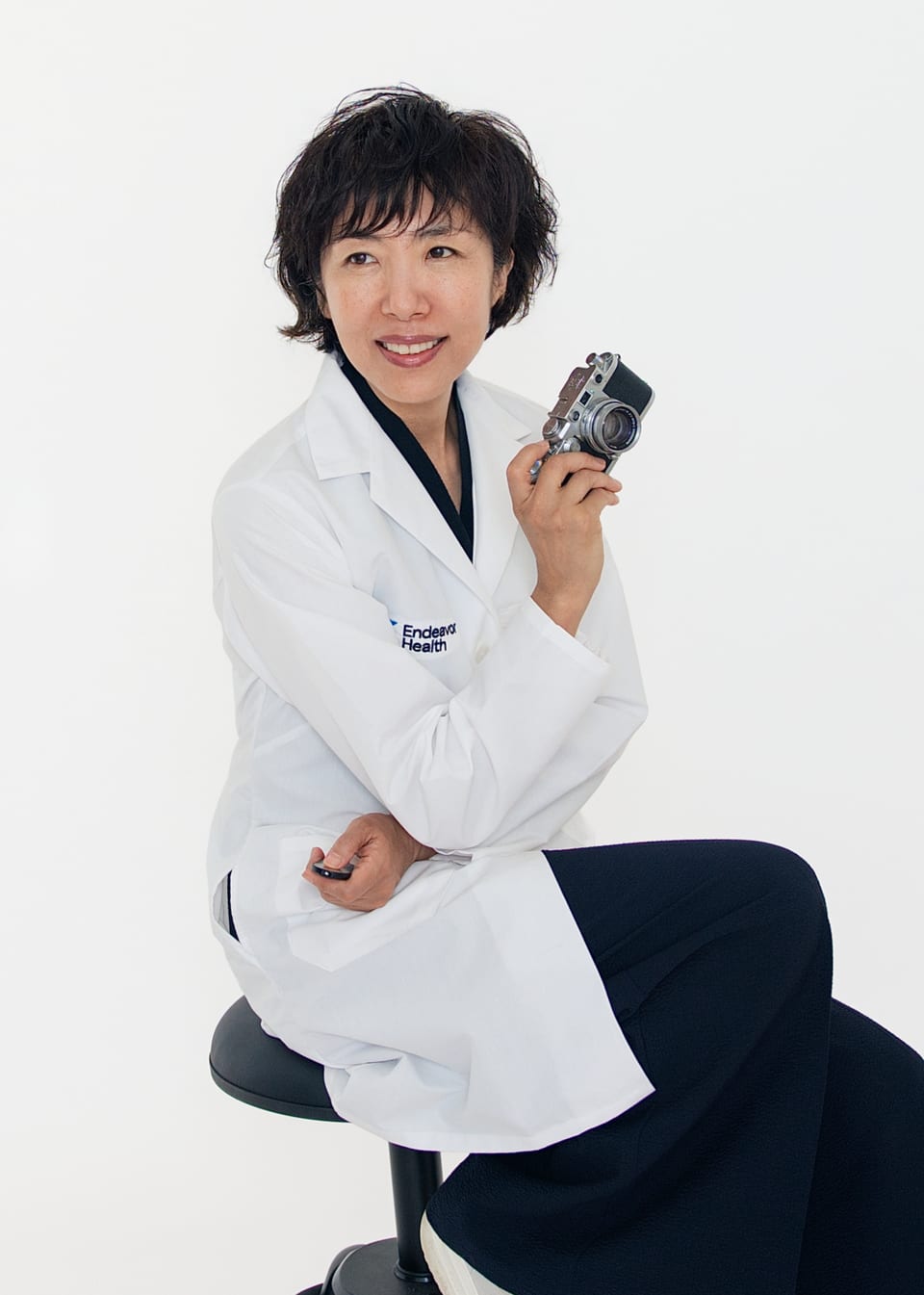 Kay Choi in a hospital white coat, holding a rangefinder camera and looking to the lens
